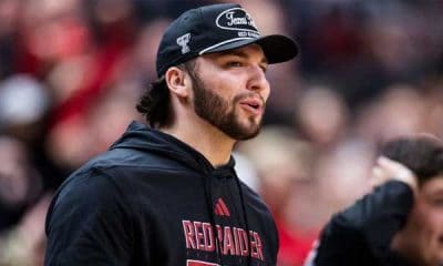 Man with a dark beard wearing a black Red Raiders hoodie and cap at a sports event, speaking or shouting to the crowd or teammates.