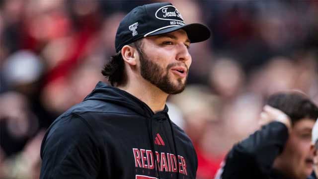 Man with a dark beard wearing a black Red Raiders hoodie and cap at a sports event, speaking or shouting to the crowd or teammates.